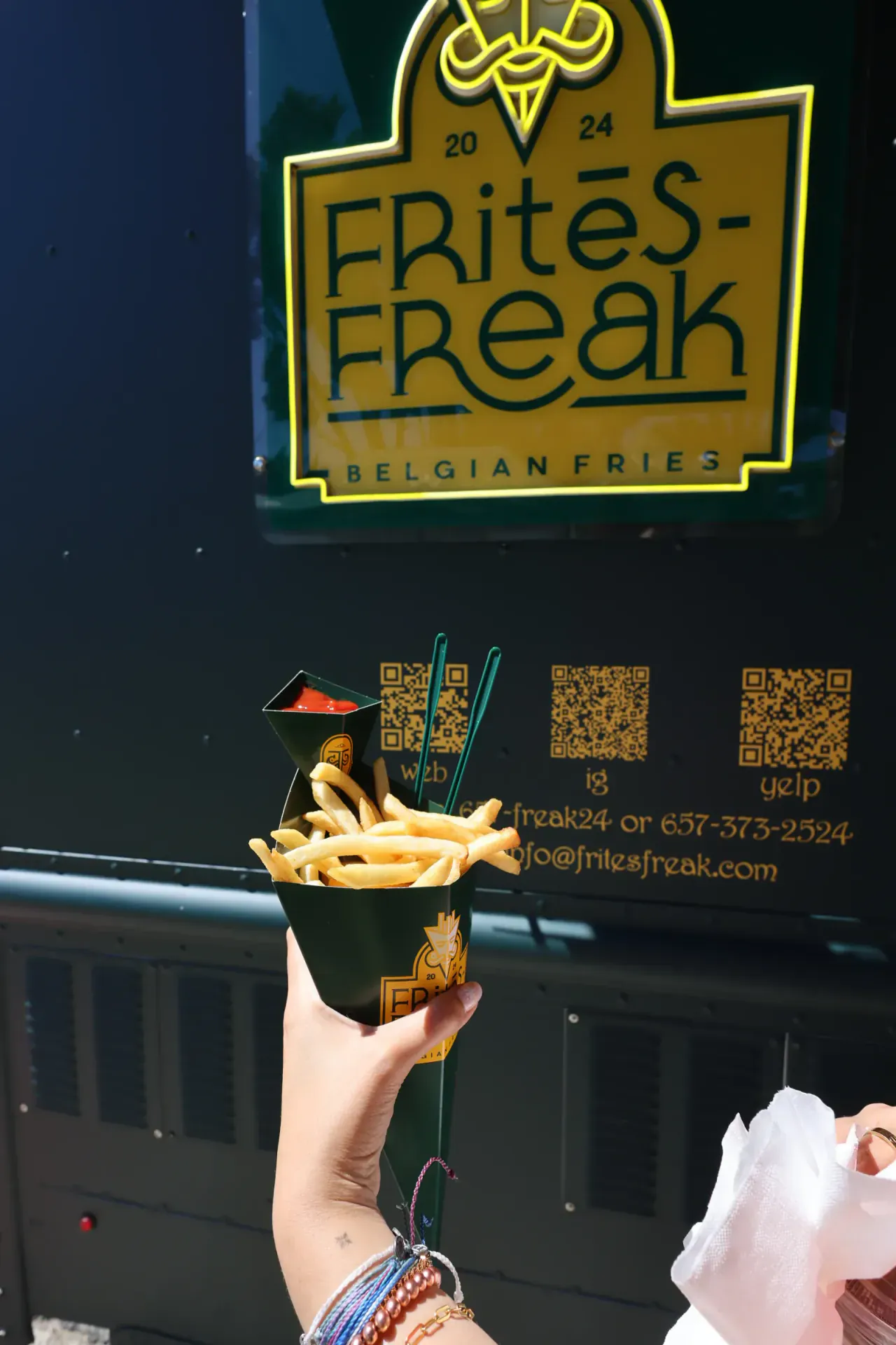 A guest holding a cone of fries and ketchup up to the truck's FRitēS-FReaK sign