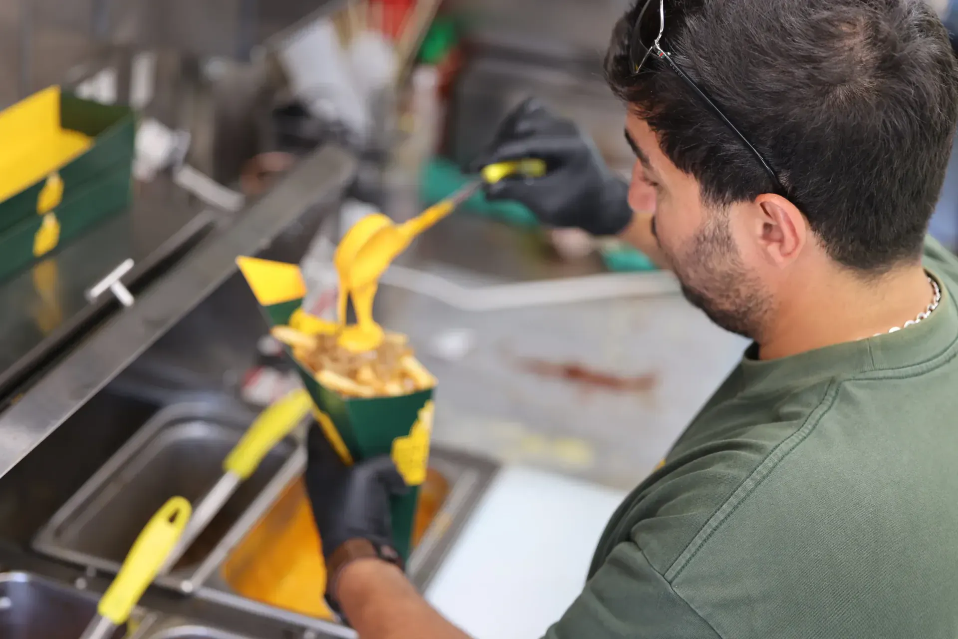A FRitēS-FReaK cook pouring warm cheese sauce over a loaded cone of hand-cut fries